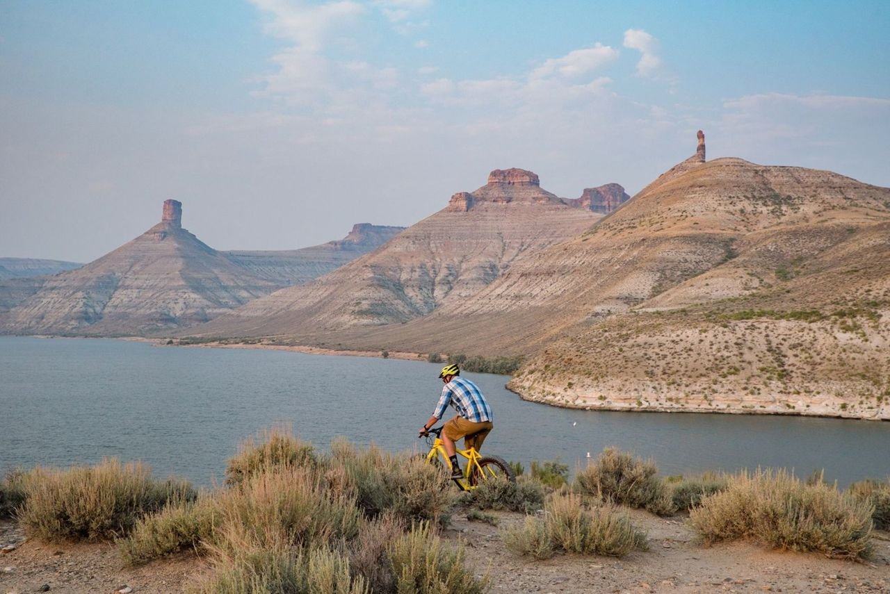 A mountain biker rides along a ridge overlooking the Flaming Gorge Reservoir with a view of Big Firehole Canyon in the distance that includes tall pinnacled buttes North and South Chimney Rock.