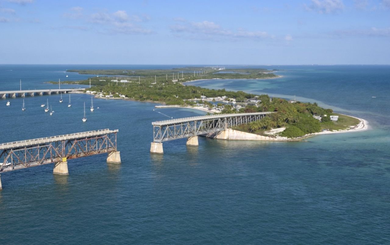 The aerial view of Bahia Honda State Park, FL, reveals the span of white, sandy beach on the Atlantic side-winding around the historic railroad bridge and sailboats moored into the bay, with the modern Florida Keys Scenic Byway and the Gulf of Mexico to the north.