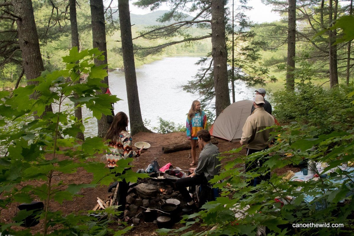 Campers cooking over a campfire. Tent visible on the site on a bluff overlooking the East Branch of the Penobscot River, Maine